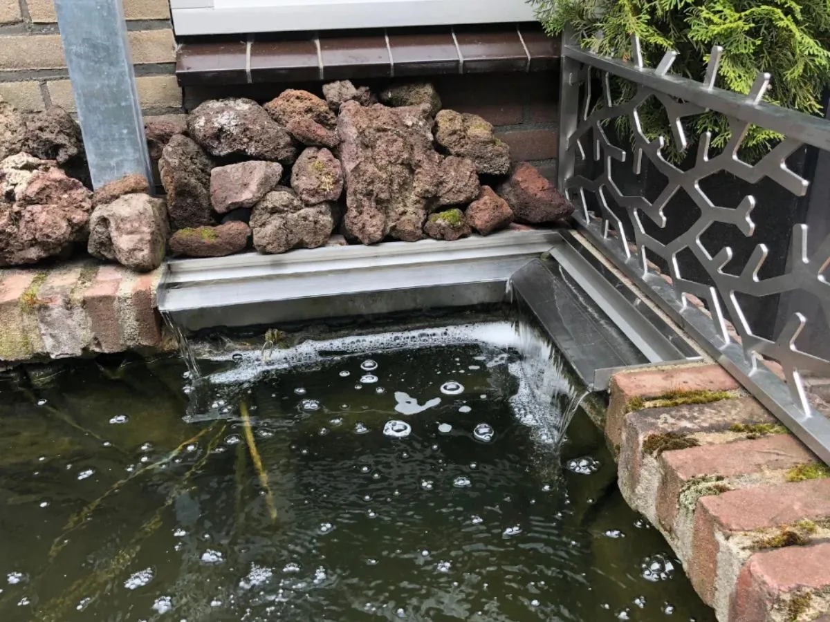 Compacte RVS waterval ingebouwd in een vijverrand van baksteen en natuursteen, waarbij het water rustig in de vijver stroomt voor een natuurlijke uitstraling.