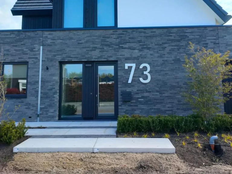 Modern RVS huisnummer 73 op een donkere grijze bakstenen gevel, duidelijk zichtbaar naast de voordeur